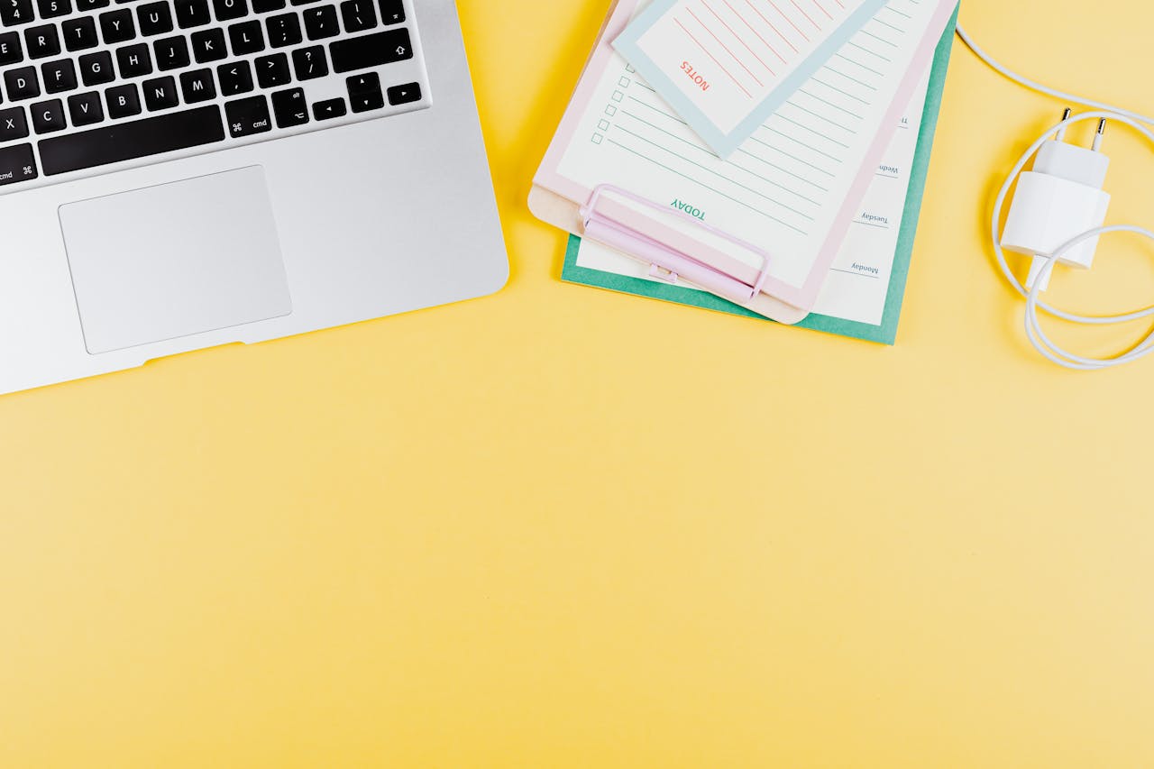 Modern minimalist workspace with a laptop, clipboard, and charger on a vibrant yellow background.